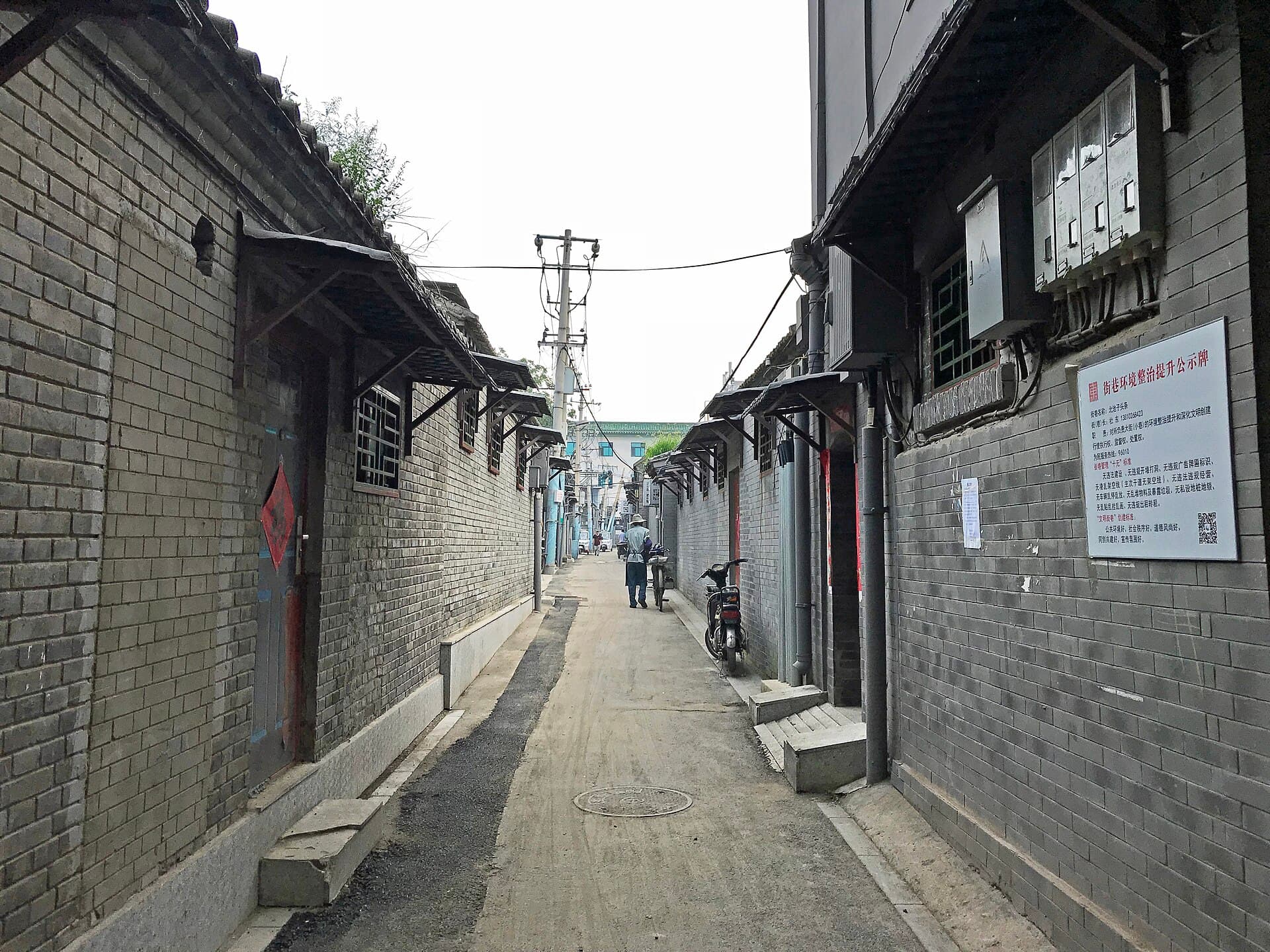 A narrow Beijing hutong lane between grey brick walls, with bicycles parked and trees leaning over