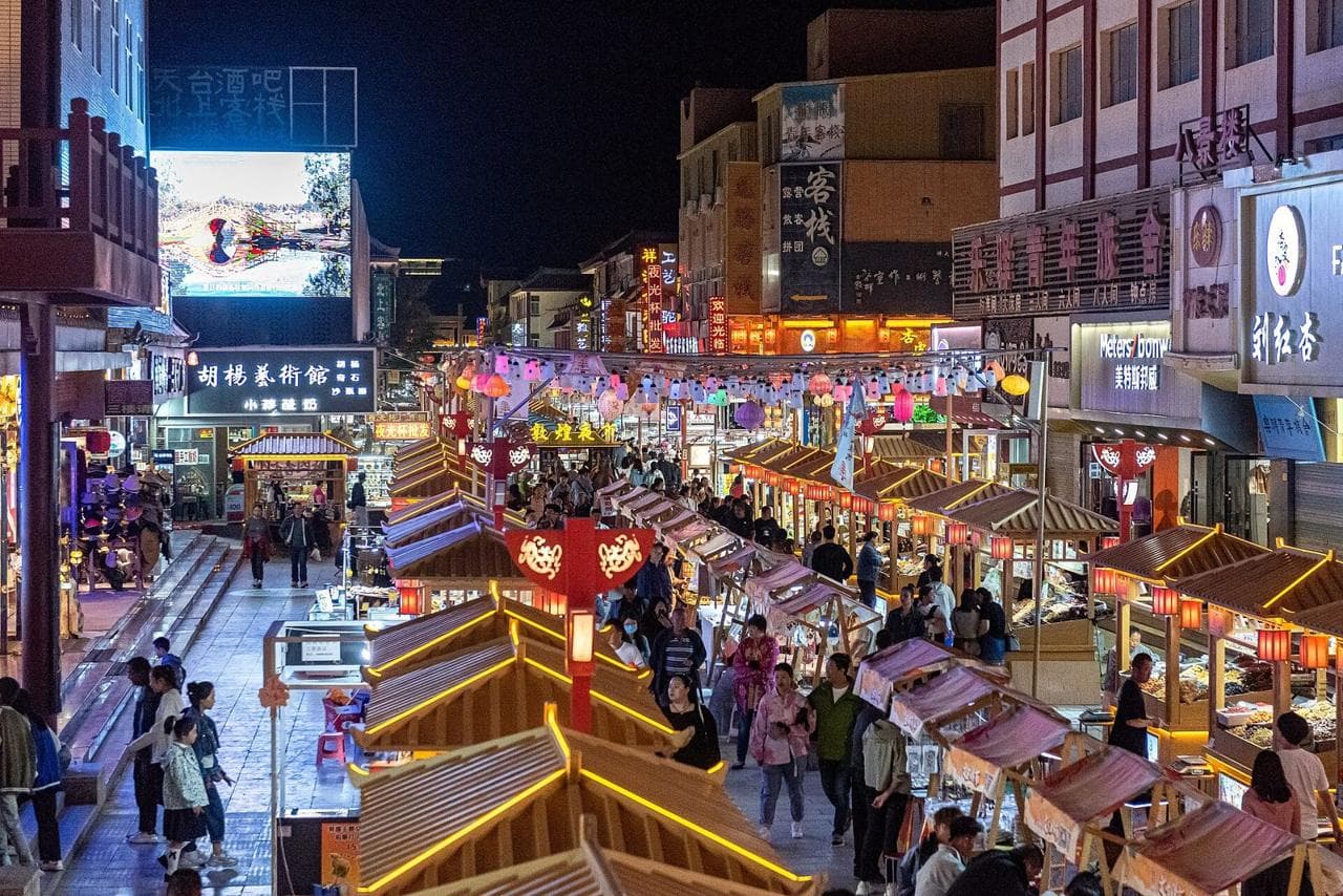 Crowds walking between rows of lit food stalls with curved roofs at the Dunhuang night market