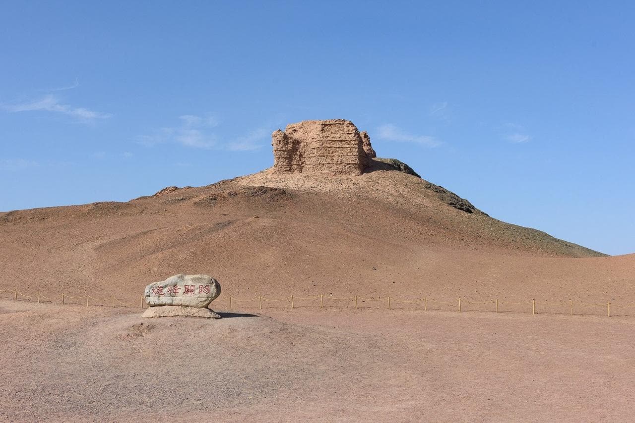 A crumbling earthen beacon tower on top of a bare desert hill, with a carved stone at its foot