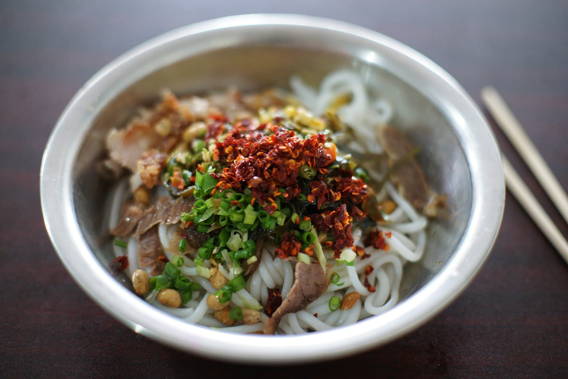 A steel bowl of Guilin rice noodles topped with sliced meat, pickles, peanuts and chilli