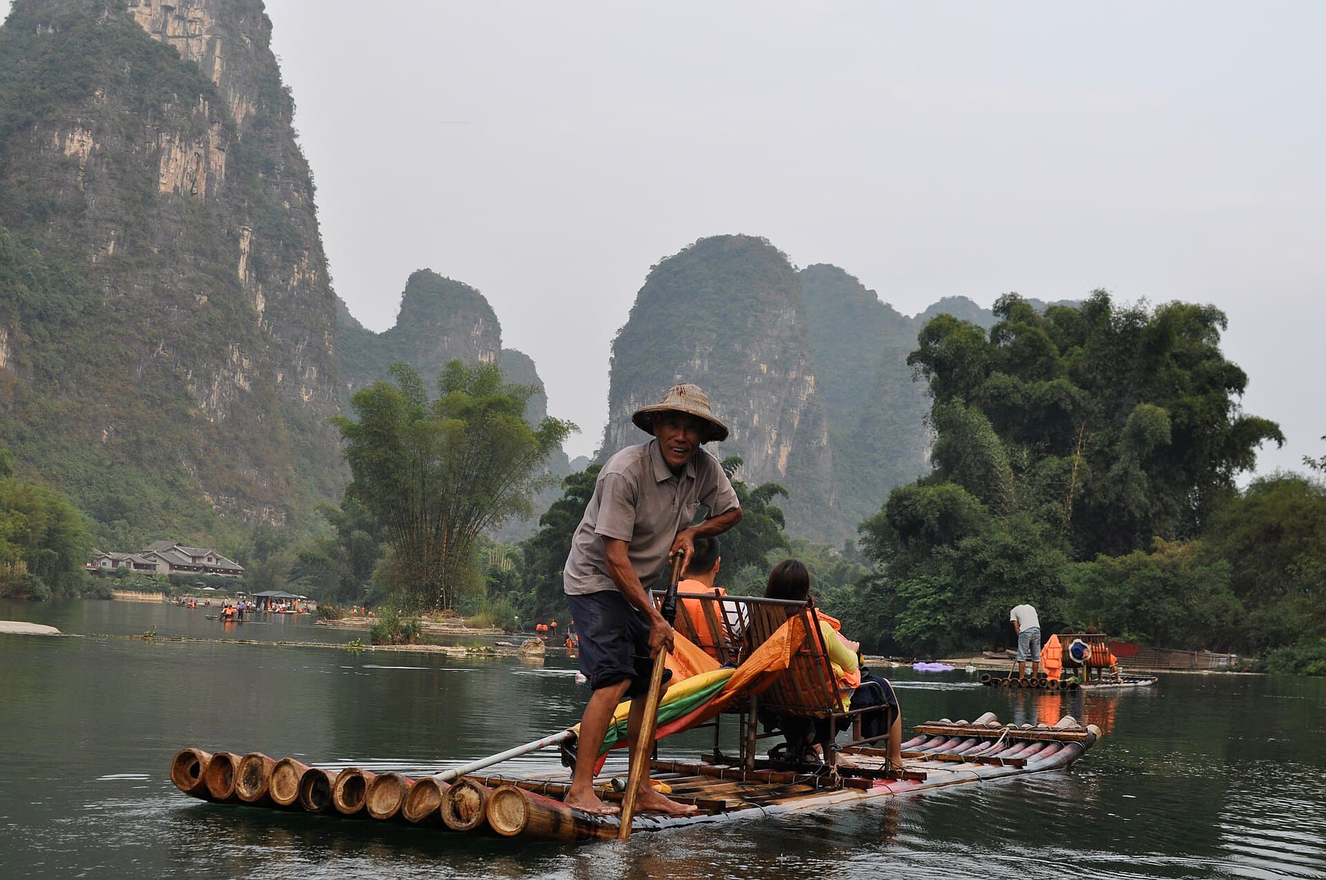 A boatman standing on a bamboo raft poling passengers along the Yulong River under karst hills