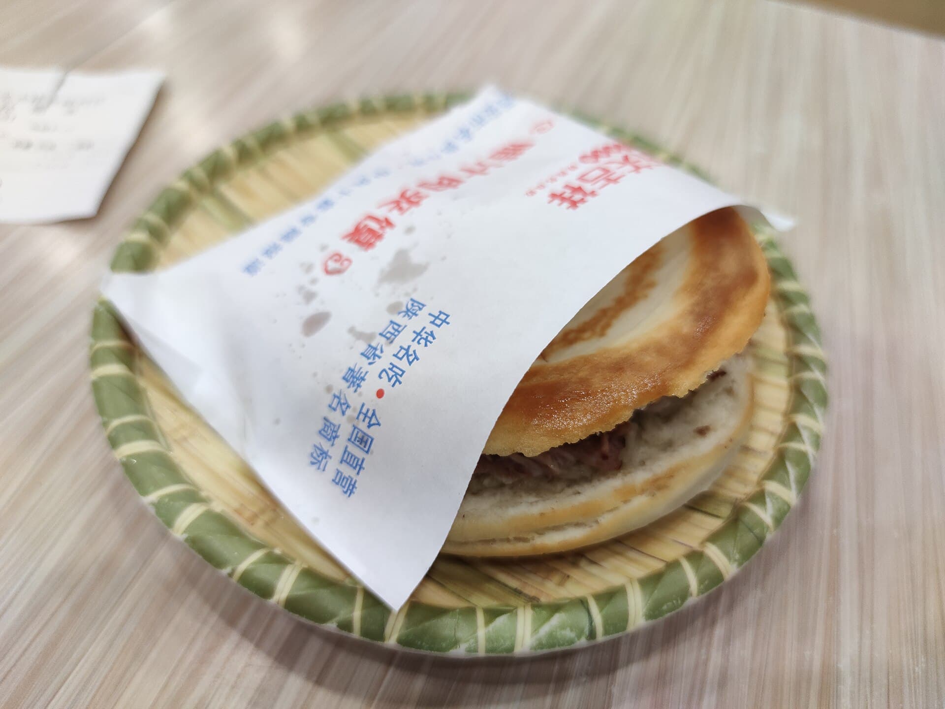 A roujiamo, a crisp flatbread stuffed with chopped braised pork, in a paper sleeve on a small plate