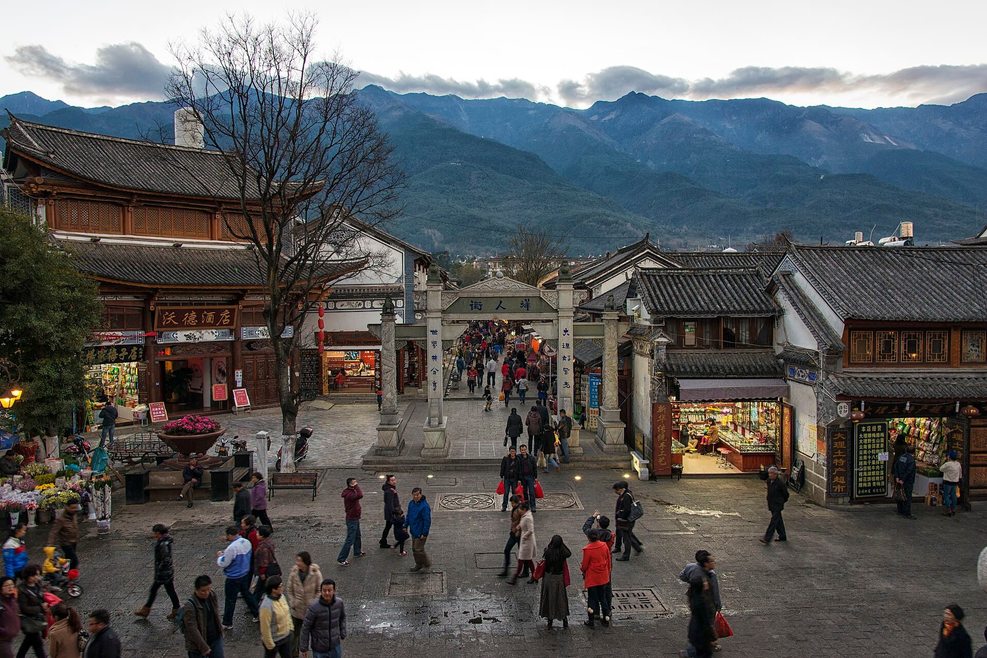 A stone-paved street in Dali Old Town with grey-tiled Bai houses, a gateway, and the Cangshan mountains behind