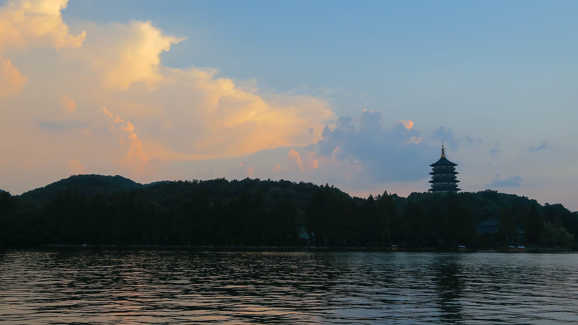 Leifeng Pagoda on a wooded hill across West Lake at dusk, with orange clouds above