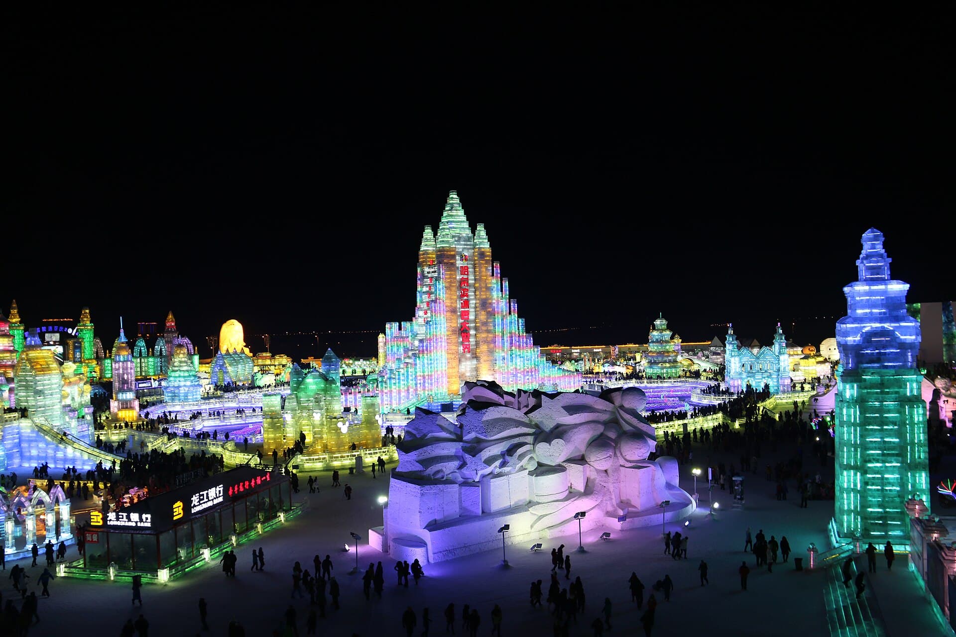 Towers and castles built from blocks of ice, lit from inside in blue, green and orange, with crowds walking between them at night